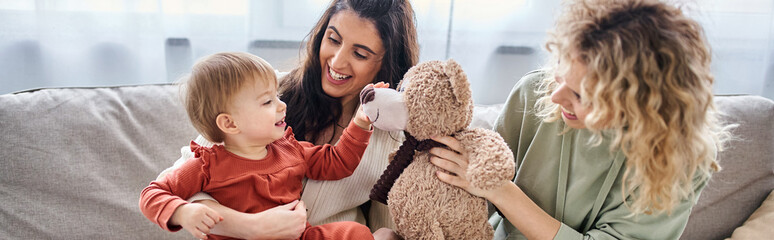 jolly lesbian couple with their toddler daughter and teddy bear on sofa, family concept, banner