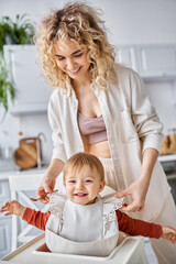 joyous beautiful blonde woman putting bib on her pretty jolly baby girl before breakfast, family