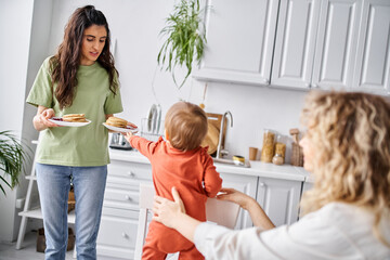 joyful brunette woman sharing fresh pancakes with her blurred blonde partner and their baby girl