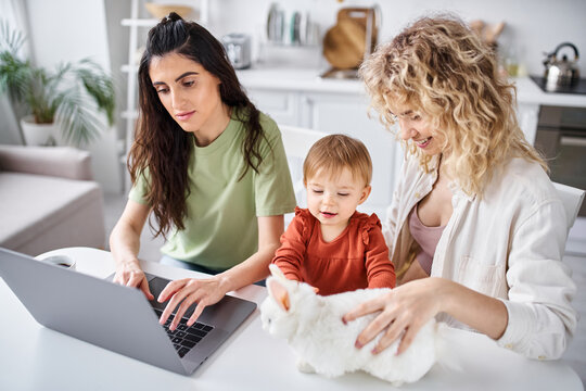 positive lesbian couple in homewear watching movies with their baby girl on laptop, family concept