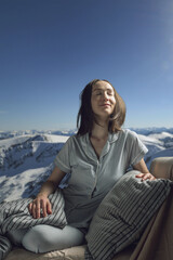 Fototapeta na wymiar A young beautiful woman in pajamas with her eyes closed in bed against the background of a lake and snowy mountains. A cozy, peaceful atmosphere of nature