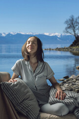 Fototapeta na wymiar A young beautiful woman in pajamas with her eyes closed in bed against the background of a lake and snowy mountains. A cozy, peaceful atmosphere of nature