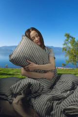 A young beautiful woman in pajamas with a pillow with her eyes closed in bed against the background of a lake and snowy mountains. A cozy, peaceful atmosphere of nature