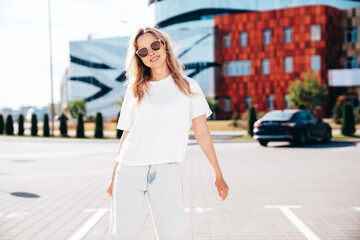 Naklejka premium Portrait of beautiful smiling model in sunglasses. Female dressed in summer hipster white T-shirt and jeans. Posing near wall in the street. Funny and positive woman having fun outdoors