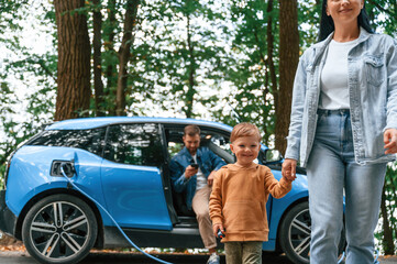 Walking forward. Mother, father and little son are waiting for electric car to charge outdoors