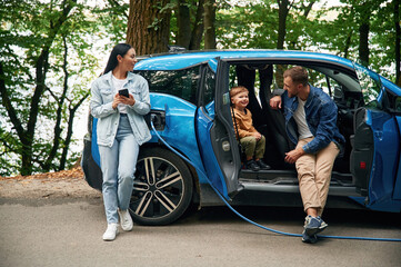 Side of the car, having fun. Mother, father and little son are waiting for electric car to charge...