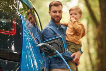 Close up view of wire in the socket. Father with his little son are waiting for electric car to charge - obrazy, fototapety, plakaty