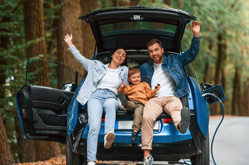 Sitting on the back of a vehicle. Mother, father and little son are waiting for electric car to...