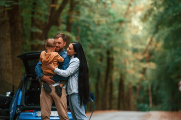 Happy family. Mother, father and little son are waiting for electric car to charge outdoors - obrazy, fototapety, plakaty