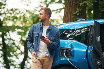 Standing and holding smartphone. Handsome man is waiting for his electric car to charge, outdoors