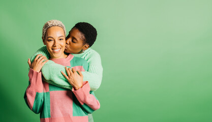 Two young cheerful women wearing green clothes embracing and kissing in a studio with a green background.