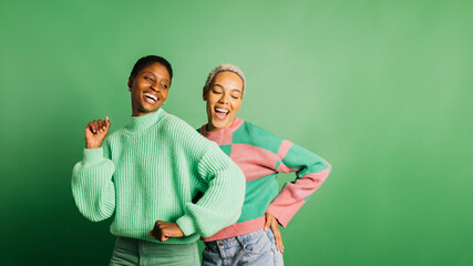Two young cheerful women wearing green clothes and dancing in a studio with a green background.