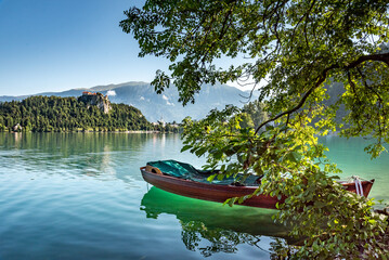 Landscape of Slovenia. A view of Lake Bled. In the foreground a boat is moored under a tree. In the background, we can see The Church  of the Assumption of Mary - obrazy, fototapety, plakaty