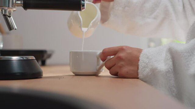 Woman adds milk to coffee in kitchen closeup. Lady in soft robe adds milky foam from jug to coffee mug enjoying start of weekend. Concept of perfect morning