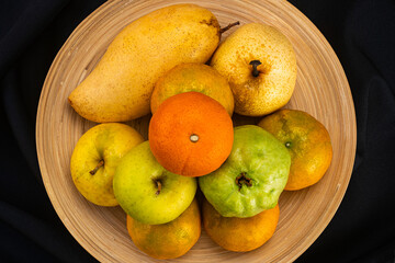 Top view pile of ripe fresh guava frui, red tomato, oranges, pear and apples in bamboo plate on black background.