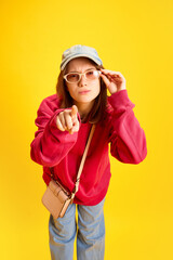 Funny girl in fashion outfit holding sunglasses and confusing looking and pointing to camera against yellow studio background. Concept of trends and style, traveling, culture, fun and joy.