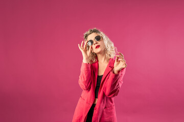 Portrait of a happy young hipster woman in a pink jacket isolated on a pink background leaning on a high chair with glasses