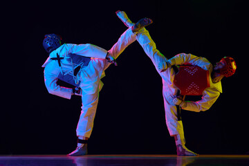 Two men in kimono and helmets practicing taekwondo, training, fighting against black background in...
