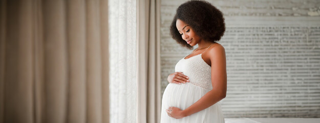 Expectant Mother Embracing Her Belly by the Window Light. A serene pregnant woman in a white dress lovingly holds her belly by a window, with gentle light caressing her figure