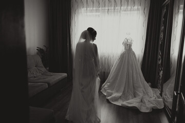 A happy bride is preparing for her luxurious wedding in a hotel room, with a wedding dress on a...