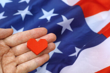 A person holding a red origami heart above the flag of the USA. Close up.
