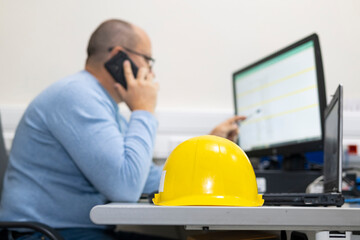 Yellow construction helmet on a work desk in the office. In the background, a man is working at a...