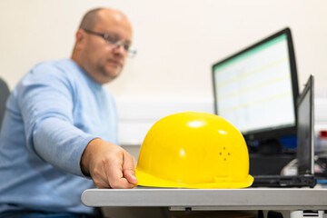 Yellow construction helmet on a work desk in the office. In the background, a man is working at a...