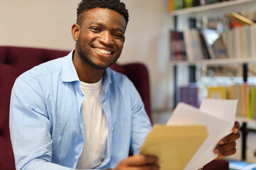 A happy young adult, whether a student or businessman, reading documents and using a computer in a modern office or university setting.