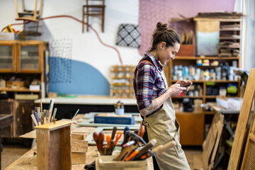 Portrait of young female carpenter using mobile phone in her workshop - obrazy, fototapety, plakaty