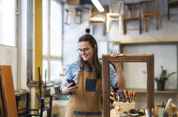 Fototapeta na wymiar Portrait of young female carpenter using mobile phone in her workshop