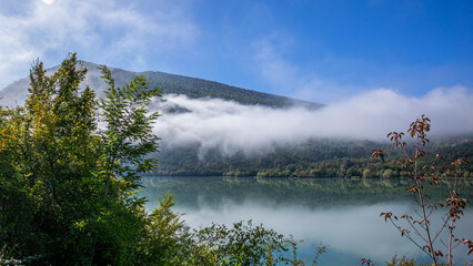 A serene landscape featuring a still lake with a rolling fog over forested hills, suggestive of...
