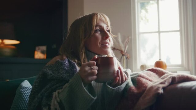 Woman wearing cosy jumper and blanket sitting on sofa at home drinking hot drink from cup - shot in slow motion