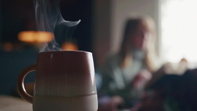 Close up of cup of steaming hot drink at home with woman reading book in background - shot in slow motion
