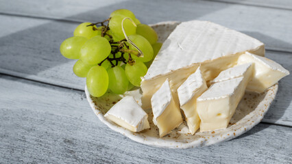 Camembert Brie cheese with grapes in a handmade clay craft plate on a blue wooden background. Side...