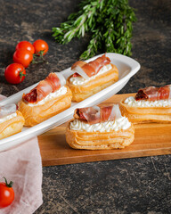 Gourmet eclair with cream filling and prosciutto on a white plate, accompanied by fresh herbs and...