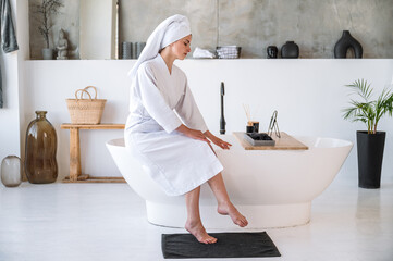 Young woman in white bathrobe sitting on bathtub in modern bathroom