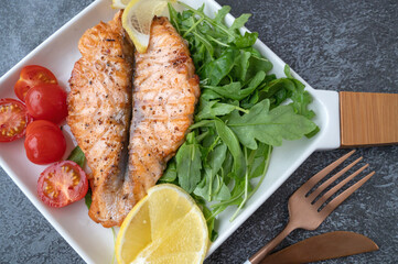 Grilled Butterfly Salmon Steak with cherry tomatoes and arugula salad on a white square plate. Close-up.