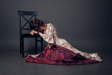 Young beautiful sad crying woman in medieval style dress sitting on the floor near chair - obrazy, fototapety, plakaty