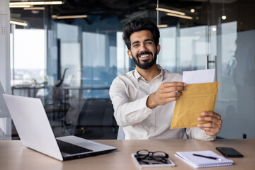 Portrait of a happy young Indian man working in the office, sitting at the desk and opening the received envelope with a letter and documents, smiling at the camera - obrazy, fototapety, plakaty