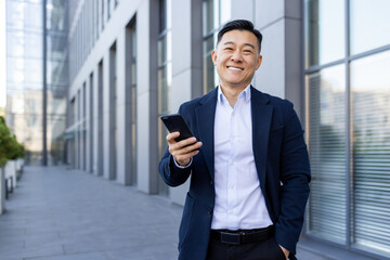 Portrait of a young Asian male businessman standing smiling on the street near an office center,...