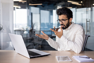 Serious young Indian male businessman sitting in the office at the desk and talking emotionally on the phone through the loudspeaker, using voice search and recording the conversation - obrazy, fototapety, plakaty