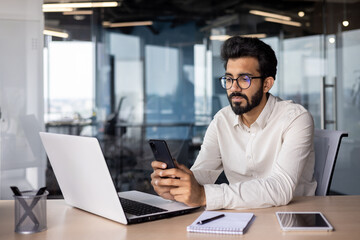 Serious Muslim young man working in office, sitting at desk and using phone, texting, checking mail...