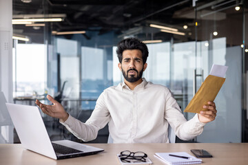 Portrait of a sad and worried young Indian man sitting in the office at the desk, holding an envelope with a letter and documents and spreading his hands, looking seriously at the camera - obrazy, fototapety, plakaty