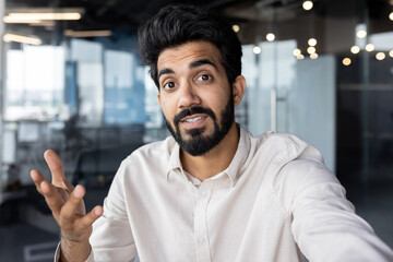 Close-up photo of a young Indian man working in the office and talking on a video call, gesturing with his hand, holding a camera - obrazy, fototapety, plakaty