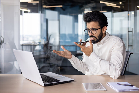 Serious young Indian male businessman sitting in the office at the desk and talking emotionally on the phone through the loudspeaker, using voice search and recording the conversation - Powered by Adobe