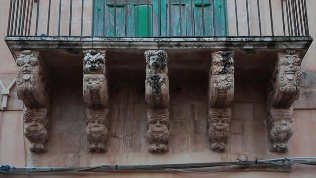 noto sicily italy statue work of faces next to each other beneath underneath balcony railing shot from straight on angle beneath with tilt up to green doors