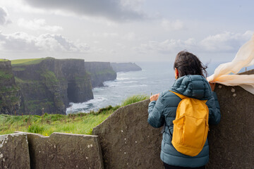 Woman on her back with backpack enjoying a splendid view from the top of the Mother's cliff in Ireland - obrazy, fototapety, plakaty
