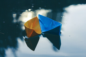 two paper boats, yellow and blue, float in the pond. The light shimmers beautifully in the water. The boat is floating side by side, like a couple in love at sunset. High quality photo