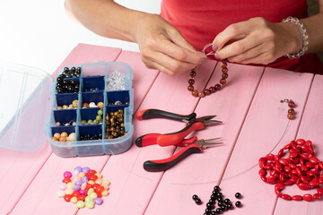 Obraz na płótnie Canvas Jewelry making. Making a bracelet of colorful beads. Female hands with a tool on a pink wooden table. Selective focus..