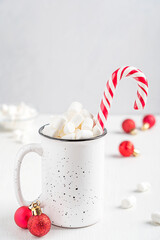 Homemade sweet hot chocolate or warm cocoa drink with marshmallow topping and striped candy cane served in mug or cup on table with christmas baubles